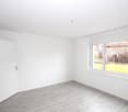 Empty room with white walls, light wooden flooring, and large windows showing a view of a green area outside.