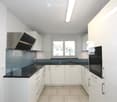 Modern kitchen with white cabinets, black countertops, and stainless steel appliances including a sink, oven, and range hood. The space is well-lit with natural light from a window.