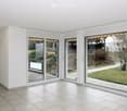 Bright, empty room with large sliding glass doors leading to a garden, white tiled floor, white walls, and a single ceiling light fixture.