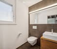 Modern bathroom with a white toilet, a wooden sink cabinet, a large mirror with built-in lighting, and a window with blinds.