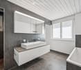 Modern bathroom with a white sink, two faucets, a bathtub, and a large mirrored cabinet above the sink. The room features dark grey tiles on the walls and floor, with large windows allowing natural light to enter.