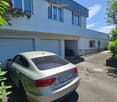 Modern two-story house with white exterior, large garage, and parking area. A silver Audi is parked in front, with bicycles stored in the garage. Greenery and a small garden are visible on the left side.