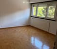 Empty room with wooden parquet flooring, white walls, large windows with blinds, radiator, and a hanging light fixture.