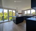 Modern kitchen with light wooden flooring, dark island, and large windows offering a scenic view.