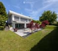 Modern white house with a spacious garden, featuring a stone patio, umbrella, fire pit, and various trees including red maples and evergreens.