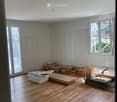 Empty room with wooden flooring, white walls, and large windows. Various construction materials and boxes are scattered on the floor.