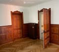 Empty room with wooden herringbone flooring, white walls, and two wooden doors with ornate carvings. A radiator is visible against one wall.