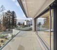 Modern balcony with large glass sliding doors, concrete floor, glass railing, and scenic view of trees and distant mountains.