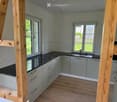Modern kitchen with grey cabinets, black countertops, and wooden flooring. Features a sink and ample natural light from large windows.