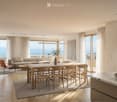 Modern living and dining area with a sofa, coffee table, dining table with chairs, bookshelf, and a kitchen island with a sink and bowl of fruit. Large windows offer a scenic view.