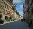 A picturesque European street lined with multi-story buildings, featuring red shutters, flags, potted plants, and a few parked cars.