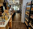 A well-lit store with wooden shelves displaying various bottles and jars of food products, wooden tables, and hanging handbags.