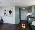 Modern open-plan living and kitchen area with wooden flooring, white walls, and large windows. Features a green sofa, a guitar, framed artwork, a kitchen counter with a toaster, and built-in cabinets.
