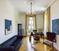 Elegant living room with wooden flooring, large windows, balcony access, a leather sofa, coffee table, desk, and decorative artwork.