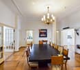 Elegant dining room with a long dark wooden dining table surrounded by matching chairs, a chandelier hanging from the ceiling, and a colorful painting on the wall. The room features light-colored walls, wooden flooring, and large glass doors leading to an