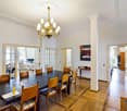 Elegant dining room with a large wooden dining table surrounded by upholstered chairs, a chandelier hanging from the ceiling, and a painting on the wall. The room features wooden flooring and opens to a living area with a sofa and television.