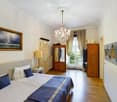 Elegant bedroom with a large bed, nightstand, lamp, wardrobe, chandelier, and artwork on the walls. The room features wooden flooring and large windows with curtains.