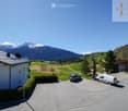 A modern house with a grey metal roof and white walls, situated in a picturesque valley with snow-capped mountains in the background. The driveway features a parked car and a van, with surrounding greenery and other houses visible in the distance.