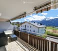 Balcony with a view of mountains and a village, featuring a sofa, a table, and a chair.