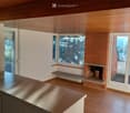 Empty room with wooden flooring, white walls, and a brick fireplace with a wooden bench. Large windows and sliding glass doors provide natural light and access to the outdoors.
