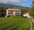 Large apartment building in the mountains, beige color, several windows and balconies, green area with trees in front