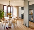 Modern dining area with a round white table, four wooden chairs, a vase with yellow flowers, and a bowl of fruit. The room features light wooden flooring, grey kitchen cabinets, a sink, and a balcony with a view.