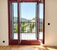 Empty room with wooden flooring, large open windows leading to a balcony with a city view.