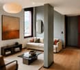 Modern living room with light-colored sofa, coffee table, side table, and artwork on the wall. Features wooden flooring, a large window, and a central column.