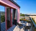 Balcony with wooden decking, black metal table and two chairs, open sliding doors leading to interior, cityscape view.