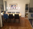 Dining area with a wooden table, six chairs, a shelf with bottles and items, and a framed picture on the wall.
