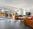 Modern living room with large glass sliding doors leading to a patio, leather sofas, a wooden coffee table, a television, and a floor lamp.