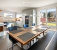 Modern kitchen with a light grey color scheme, featuring a central wooden dining table surrounded by blue chairs, a kitchen island with a countertop, and built-in cabinets with stainless steel appliances.
