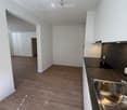 Empty kitchen with wooden flooring, white walls, and a stainless steel sink. No furniture or appliances are present.