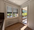 Bright room with wooden flooring, radiator, large windows with blinds, and a sliding door opening to a balcony with outdoor view.