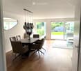 Bright dining room with wooden flooring, a black dining table surrounded by six chairs, pendant lights above, and a large mirror on the wall. A sliding glass door leads to a garden view.
