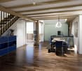 Open-plan living and dining area with wooden flooring, exposed wooden beams, modern pendant light, dining table with chairs, kitchen island, and potted plants.