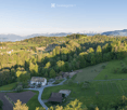 Aerial view of a rural landscape with a house, outbuildings, and surrounding fields and forests, with mountains in the background.