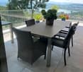 Balcony with a modern glass railing, a large table with four wicker chairs, and potted plants, overlooking a scenic lake and mountain view.