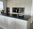 Modern kitchen with white cabinets, black countertop, stainless steel sink, and various appliances including an oven and fridge. The space is well-lit with recessed ceiling lights and features a large window with a view.