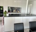 Modern kitchen with white cabinets, black countertops, and a central island. Features include a sink, two black leather chairs, a small table, and a potted plant on the counter.