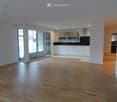 Empty room with wooden flooring, white walls, and a large glass door leading to a balcony. The kitchen area is visible in the background with white cabinets and stainless steel appliances.
