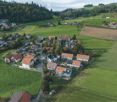 Aerial view of a rural village with multiple houses, green fields, and forested areas in the background.