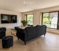 Modern living room with large windows overlooking a scenic view, featuring a dark grey sofa, ottoman, coffee table, television, and a potted plant.