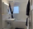 Modern bathroom with a white sink, bathtub, toilet, mirror, radiator, and a window with curtains. Towels are placed on the radiator and floor.