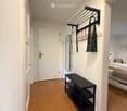 A narrow hallway with light-colored walls and carpeted floor, featuring a black bench, coat rack, and a glimpse of a bedroom with a bed and nightstand.