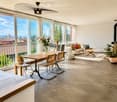 Modern living room with large windows offering a scenic view, featuring a white sofa, wooden dining table, chairs, and decorative items.
