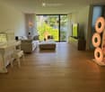 Modern living room with wooden flooring, large windows, a sofa, ottoman, piano, and decorative art pieces.