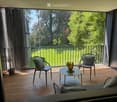 Modern balcony with wooden decking, two chairs, a small table, a sofa, and a decorative vase. Large windows offer a view of a green lawn and trees.