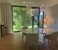 Modern dining room with a round glass table, four upholstered chairs, large windows with curtains, and decorative lighting fixtures.