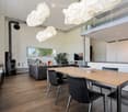 Modern open-plan living and dining area with wooden flooring, a grey sofa, a wooden dining table with black chairs, a fireplace, a television, and several potted plants. The space features large windows and modern pendant lights.